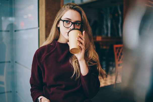 Pondering female student enjoying morning takeaway coffee after university resting indoors, pensive hipster woman wear in eyeglasses thoughtful looking aside while spending time at cozy cafeteria - Photo, image