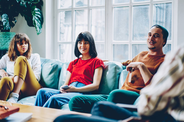 Cheerful group of young people dressed in casual wear laughing during playing game sitting on cozy couch in modern apartment.Positive male and female spending time together during friendly meeting - Photo, image