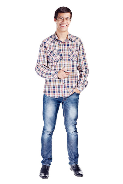 Full body portrait of smiling young man with mobile phone in hand, other hand in pocket wearing metal frame glasses, checkered shirt, blue jeans and black shoes isolated on white background - Photo, image