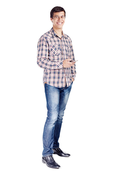 Full body portrait of smiling young man with mobile phone in hand, other hand in pocket wearing metal frame glasses, checkered shirt, blue jeans and black shoes isolated on white background - Fotó, kép