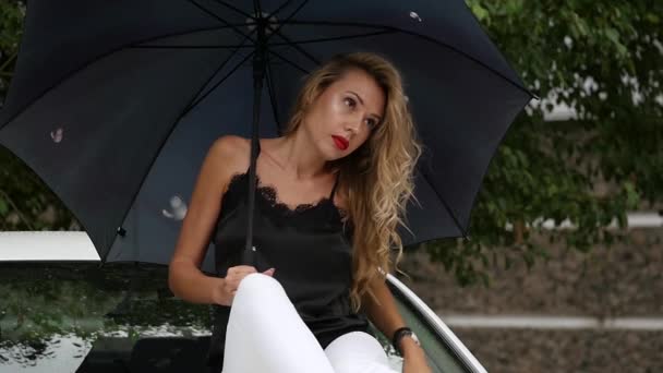 Beautiful young woman with long blonde hair sitting on the hood of the white car with umbrella in rainy summer day. slow motion - Кадри, відео