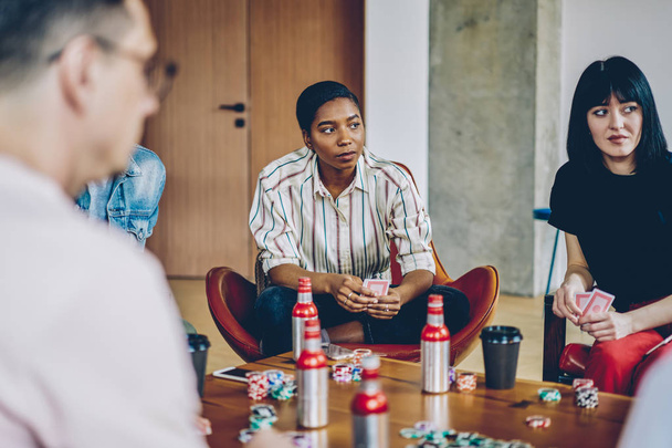 Concentrated diverse hipster guys with cards in hands playing poker at table with chips and drink in modern flat.Casual dressed young people spending free time together in apartment - Fotoğraf, Görsel