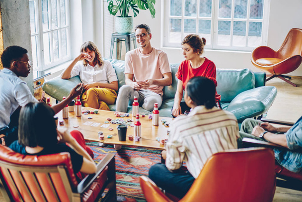 Positive diverse hipsters laughing while playing game at poker table in modern apartment.Cheerful multicultural casual dressed young men and women having fun enjoying entertainment in flat - Foto, Imagem