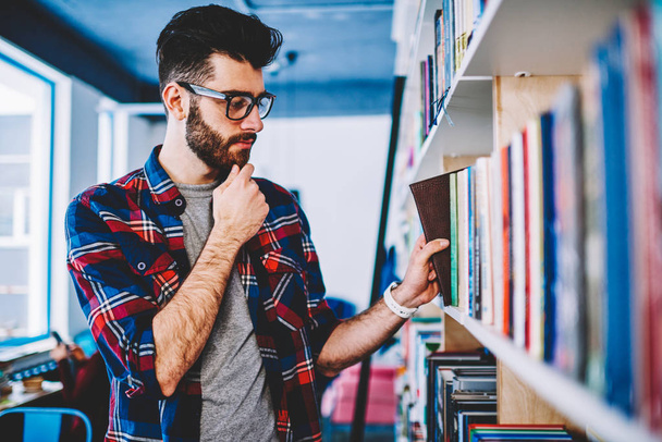 Hombre reflexivo en gafas seleccionando libro en la biblioteca de la universidad de pie cerca de los estantes, tipo hipster concentrado casual vestido recogiendo literatura y haciendo investigación en la librería favorita
 - Foto, imagen