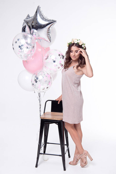 happy smiling woman with curly hairstyle and floral wreath posing near wooden chair with balloons isolated on white background - Photo, image