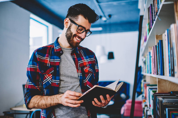 Clever smiling caucasian male in casual wear reading new interesting novel in bookstore, successful joyful man student holding positive book while standing near shelves in university library  - Foto, imagen