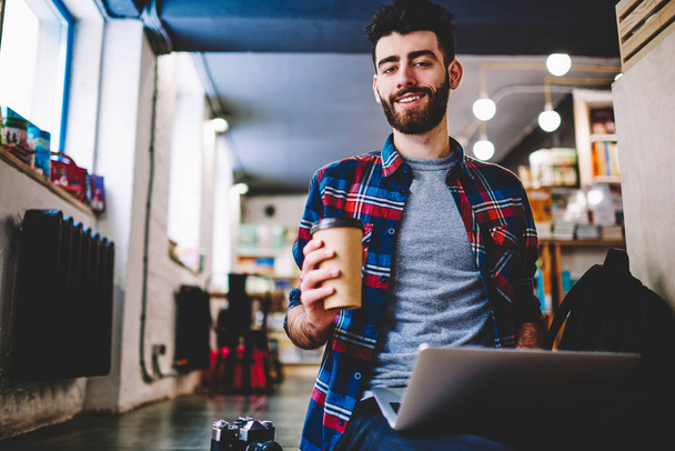Portrait of successful hipster guy enjoying morning takeaway coffee at university campus and spending time for e learning, happy male copywriter looking at camera in break of work on laptop computer - Photo, image
