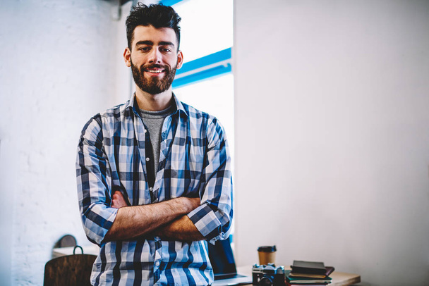Half length portrait of successful male teenager looking at camera with cute smile on face at college campus, positive caucasian bearded hipster guy wear in trendy casual look enjoying time indoors - Foto, imagen