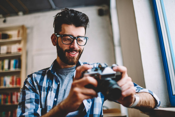 Successful hipster guy waiting friend in bookstore while checking photos at vintage camera during free time, happy stylish smiling man wear in trendy apparel holding old fashioned equipment - 写真・画像