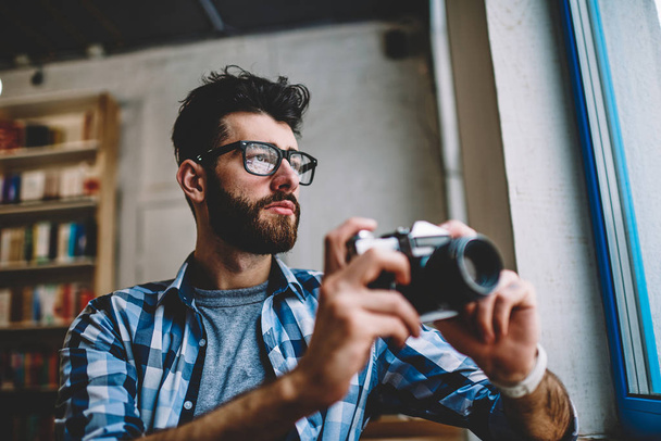 Thoughtful hipster guy wear in spectacles for vision correction looking in window after making images via retro camera, concentrated professional male photographer enjoying time and hobby indoors - Photo, Image