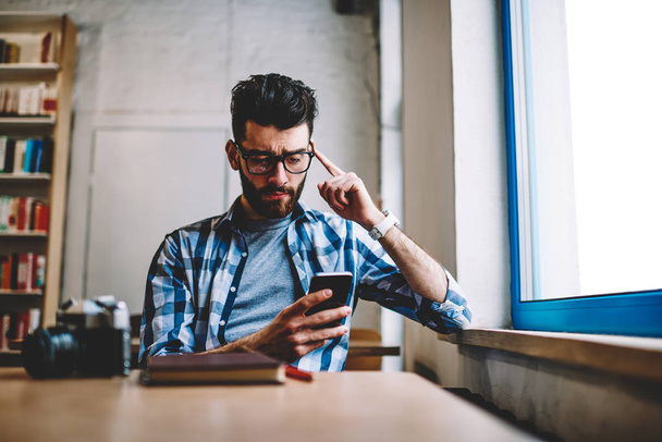 Joven hipster confundido en gafas leyendo mensaje de texto desde el trabajo usando aplicación en el teléfono móvil, concentrado blogger masculino descargando archivo en un teléfono inteligente moderno conectado a wifi en interiores
 - Foto, imagen