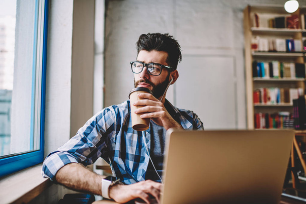 Thoughtful hipster guy enjoying new playlist via earphones drinking morning coffee sitting desktop with laptop computer at coworking space, puzzled man looking away and waiting webinar on netbook - Φωτογραφία, εικόνα