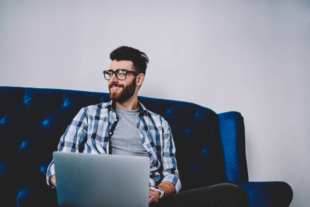 Happy caucasian blogger sitting on cozy sofa with modern laptop computer and enjoying e learning, successful hipster guy in eyewear looking aside with cute smile on face in break of working on netbook - Photo, Image