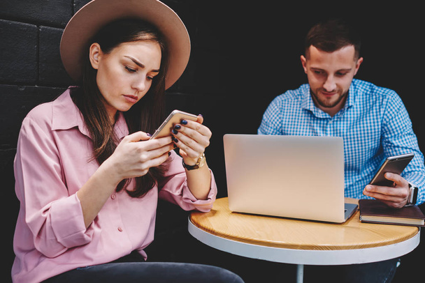 Потрясающая молодая женщина читает странные входящие уведомления на смартфоне, пользуясь бесплатным интернетом 4G, сидя с бойфрендом, который работает на фрилансе за современным ноутбуком в кафе.
 - Фото, изображение