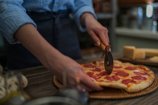 Woman cutting fresh baked homemade pizza on rustic kitchen background. Cut into slices delicious pizza with mushrooms and ham. Cheese and tomatoes on wooden table. Healthy foods, cooking concept. - Fotó, kép