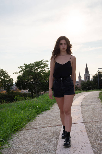 young beautiful girl walks in a city park. Young girl with long brown hair, in black tank top and jeans shorts while walking on a narrow street in Verona, Italy - Foto, Imagen