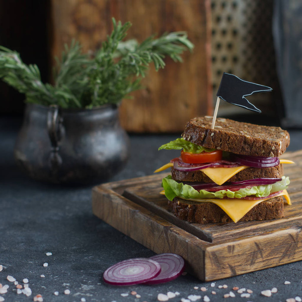 tasty sandwich made of bread, tomatoes,sausage, onion and lettuce on a wooden board and dark background, selective focus - Foto, imagen