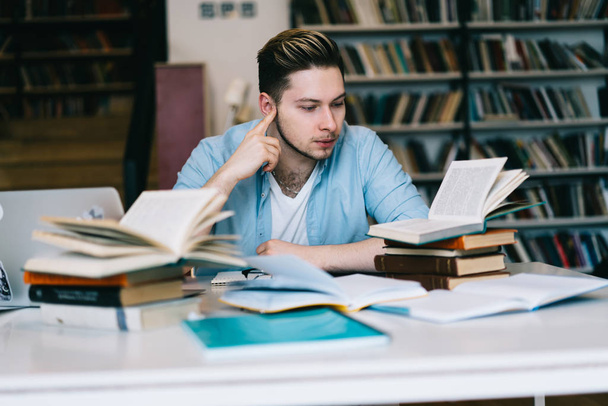 Pensive male student sitting at desktop learning information for university examination, clever hipster guy spending time on autodidact reading textbook and literature doing homework in library
 - Фото, изображение