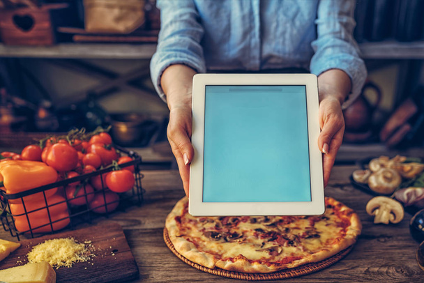 Hand holding a tablet computer with white screen. Woman hands showing empty screen of modern digital tablet. Cooking and digital everyday life online concept. Screen in focus with woman in kitchen. - Fotoğraf, Görsel