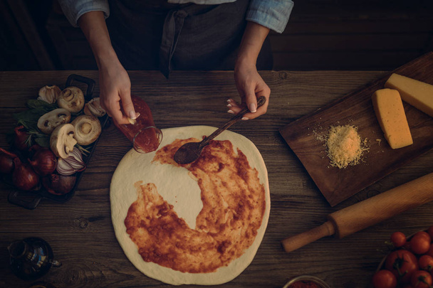 The process of making pizza. Prepare dough hand topping. Woman hand is spreading pasteurized tomato paste onto a pizza base. Food concept. Preparing traditional italian pizza. Toned image. Top view. - Photo, Image