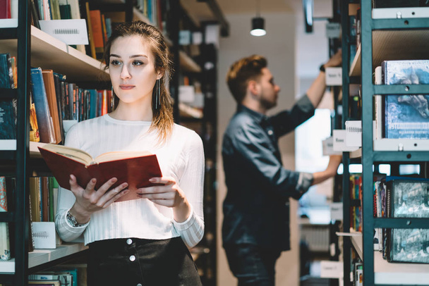 Serious female college student in casual wear pondering on literature standing near bookshelves in store,attractive young woman librarian concentrated on job holding book making order in librar - Foto, Imagem
