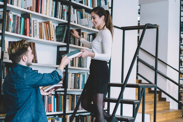  Cheerful young woman standing on ladder get textbook satisfied with help of boyfriend, female librarian giving literature to male student helping her to go down on staircase near bookshelves - 写真・画像