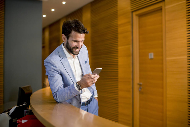 Retrato de jovem empresário em pé no escritório e usando telefone celular
 - Foto, Imagem