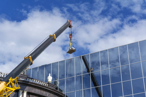 телескопическая стрела строительного крана поднимает нагрузку на зеркальную стену здания, отражающую небо
 - Фото, изображение