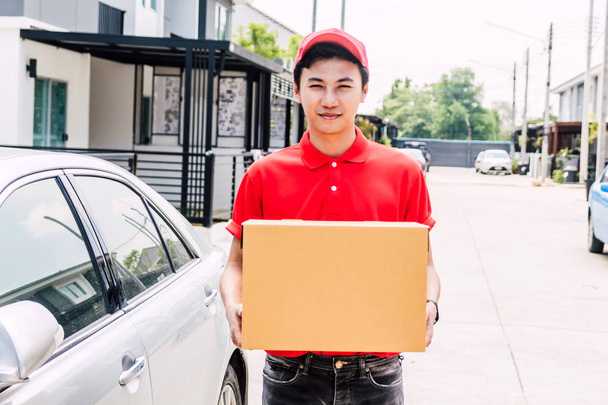 Glücklicher Zusteller in roter Uniform mit Karton, der in der Nähe des Hauses des Kunden steht. - Foto, Bild