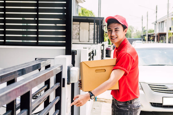 Счастливый человек в красной форме, держащий картонную коробку, стоящую возле дома клиента. Концепция курьерской службы - Фото, изображение