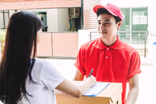 Женщина кладет подпись в буфер обмена на картонной коробке, чтобы получить пакет с доставщиком мужчина в красной униформе.
 - Фото, изображение