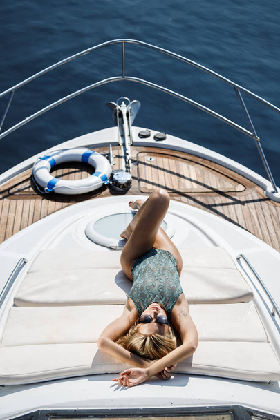 Beautiful blonde woman wearing green lingerie and black sunglasses posing to camera during travelling on luxury yacht at daytime - 写真・画像
