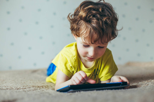 Little cute boy in a green T-shirt playing games on a tablet and watching cartoons - Fotó, kép