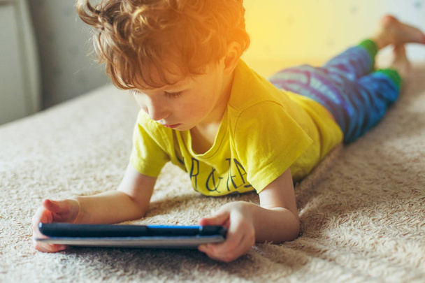 Little cute boy in a green T-shirt playing games on a tablet and watching cartoons. addiction concept - Φωτογραφία, εικόνα
