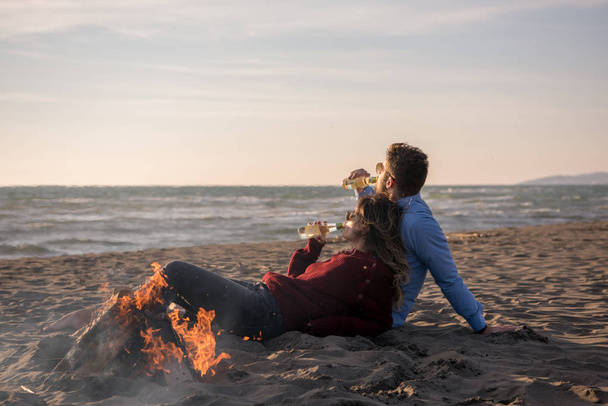 Nuori pari rentoutumassa tulen ääressä, juomassa olutta tai juomassa pullosta rannalla syyspäivänä - Valokuva, kuva