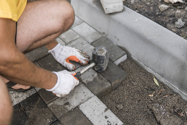 Hände eines Bauarbeiters in seinen orangefarbenen Handschuhen mit einem Hammer, der neue Pflastersteine im Außenbereich sorgfältig auf einen ebenen und geharkten Boden legt. Sandfundament. Gebäude. - Foto, Bild