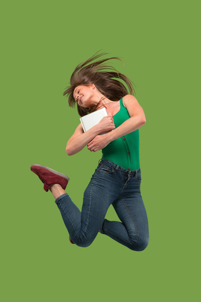 Image of young woman over green background using laptop computer or tablet gadget while jumping. - 写真・画像