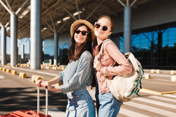 Kaunis hymyilevä tytöt aurinkolasit onnellisesti etsivät kameran tilalla matkalaukku ja reppu olkapäällä lentokentällä taustalla
 - Valokuva, kuva