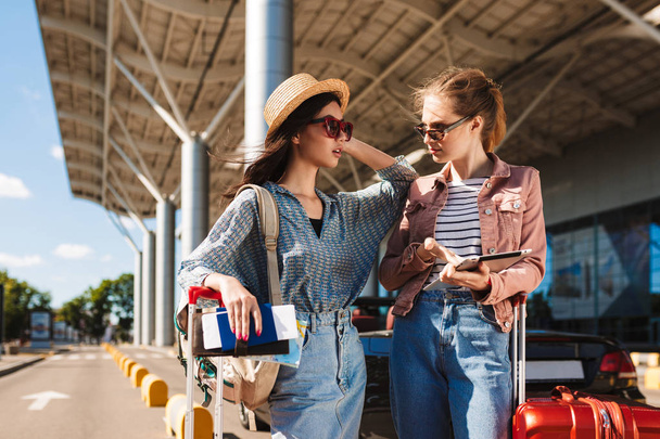 Jovens amigas em óculos de sol incrivelmente olhando uma para a outra segurando passaporte com bilhetes e tablet nas mãos com o aeroporto em segundo plano
 - Foto, Imagem