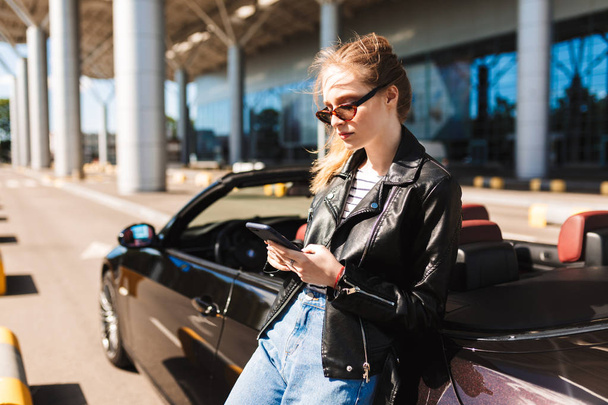 Menina bonita em óculos de sol e jaqueta de couro encostada no carro cabriolet preto pensativamente usando celular com aeroporto no fundo
 - Foto, Imagem