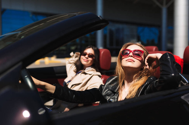 hübsches Mädchen mit Sonnenbrille schaut verträumt in die Kamera fahrendes Cabrio mit Freundin in der Nähe auf den Straßen der Stadt im Freien - Foto, Bild