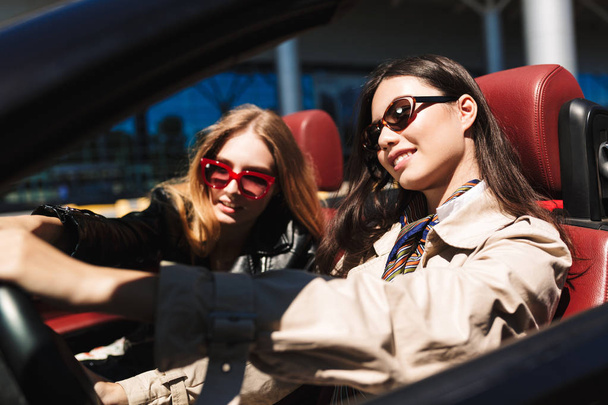 Pretty smiling girl in sunglasses happily looking aside driving cabriolet car with female friend near on city streets outdoor - Foto, imagen