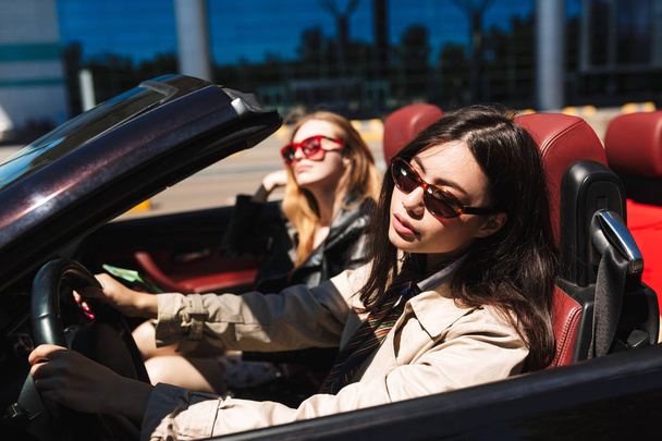 Mooi meisje in zonnebril dromerig besturen cabriolet auto met vriendin in de buurt van op straat buiten - Foto, afbeelding
