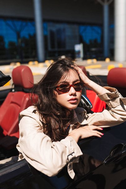 Beautiful girl in sunglasses and trench coat leaning on cabriolet car door thoughtfully looking in camera with airport on background - Photo, Image