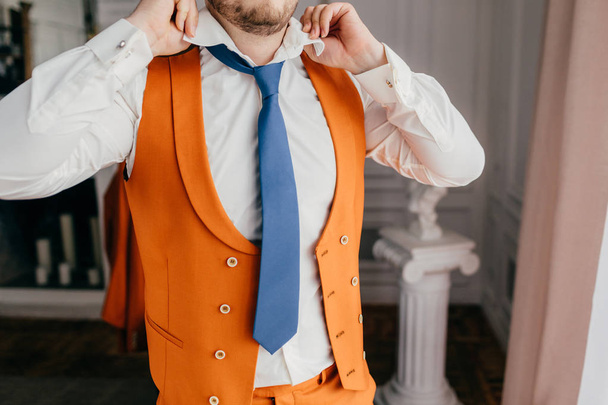 Cropped shot of unrecognizable male groom ties tie, dressed in elegant white shirt and orange jacket, prepares for special day in his life, stands indoor. Wedding, preparation, celebration concept - Photo, Image