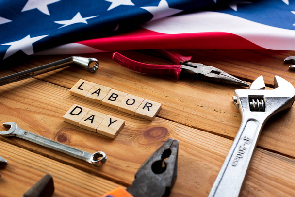 Концепция Дня труда США, первый понедельник сентября. Различные виды ключей, удобные инструменты, флаг Америки и деревянные блоки на деревянном столе
. - Фото, изображение
