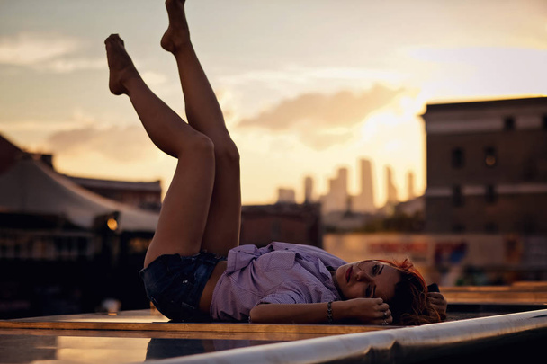 schöne Frau mit roten Haaren sitzt auf einem Dach oder einer Brücke, Sonnenuntergang Licht - Foto, Bild