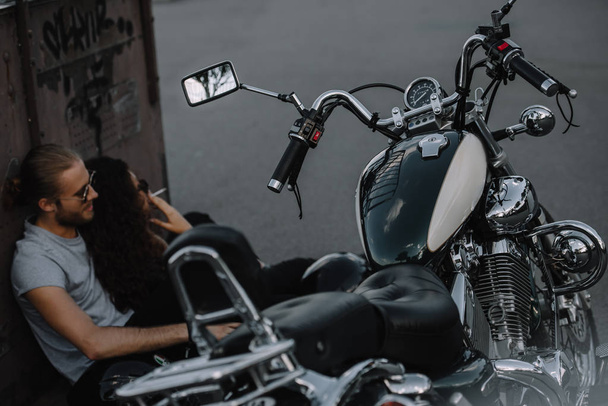 foco seletivo de motociclistas fumando e sentado no asfalto perto de motocicleta clássica
 - Foto, Imagem