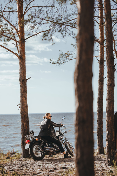 motociclista em jaqueta de couro preto sentado na motocicleta clássica perto do mar na floresta
 - Foto, Imagem