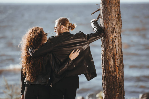 back view of couple in black leather jackets hugging and looking at each other on seashore - Photo, image